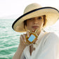 A person in a straw hat holds a Sirena - Eau de Parfum by Floris London by the sea, sunlight highlighting their features.