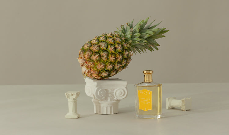 A pineapple rests on a small white pillar next to a bottle of Floris perfume and two tiny pillar ornaments.