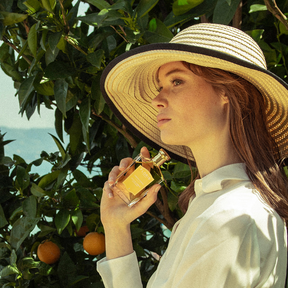 Woman in straw hat holds Floris London's Soulle Ámbar perfume near orange trees, enveloped by warm amber and fruity pineapple notes.