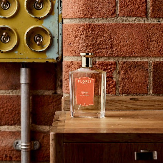 A Floris London Special No. 127 Eau de Toilette bottle rests on wood by a brick wall and vintage yellow electrical panel, evoking citrus floral notes.