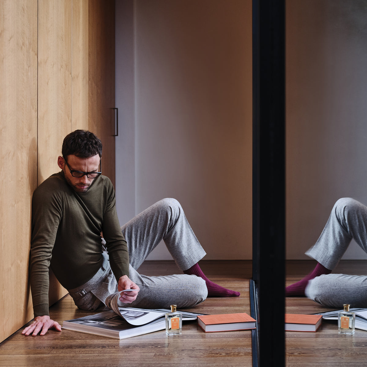 A man seated on the floor reading books is reflected in a mirror, with a bottle of Floris London's Special No. 127 - Eau de Toilette beside him.