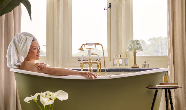 A woman relaxing in a green bathtub in front of a window, surrounded by various Floris Bath & Body products.