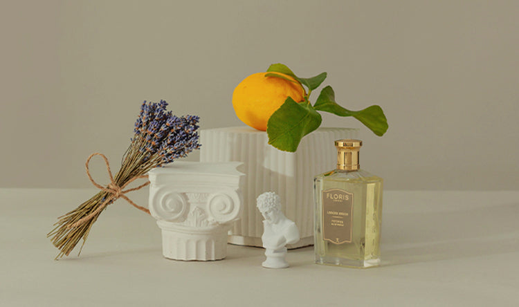 Still life with a lemon, lavender flowers, a small white bust, a perfume bottle, and classical decor on a neutral background.