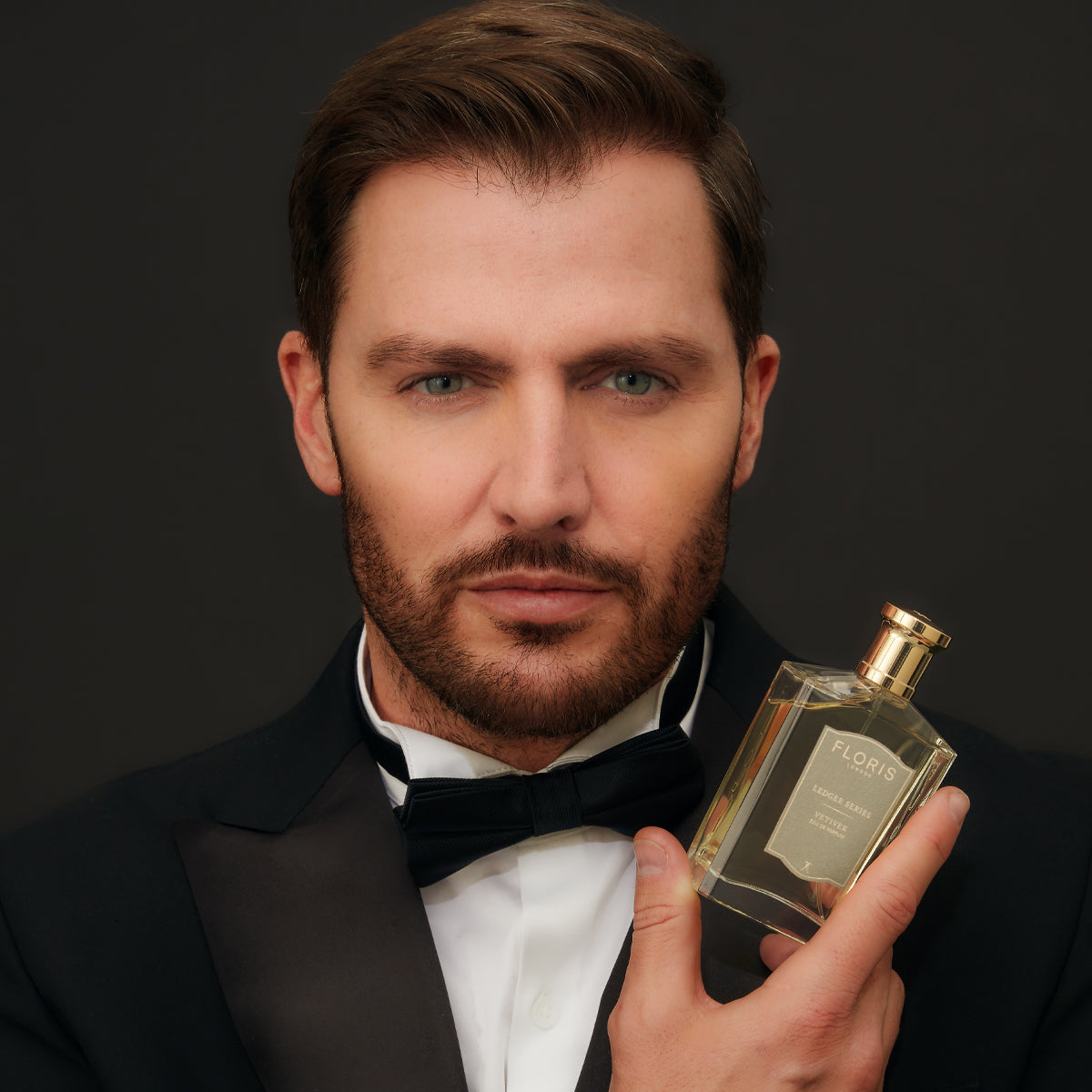 A man in a formal suit holds a Floris London Vetiver perfume bottle, radiating an earthy essence, against a dark background.