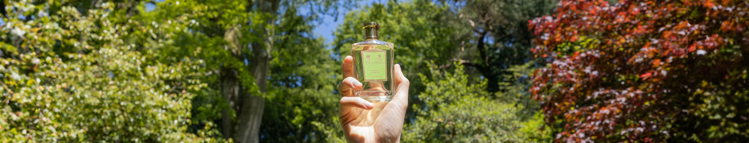 A hand holding a perfume bottle against a backdrop of lush green trees and a red-leaved tree.