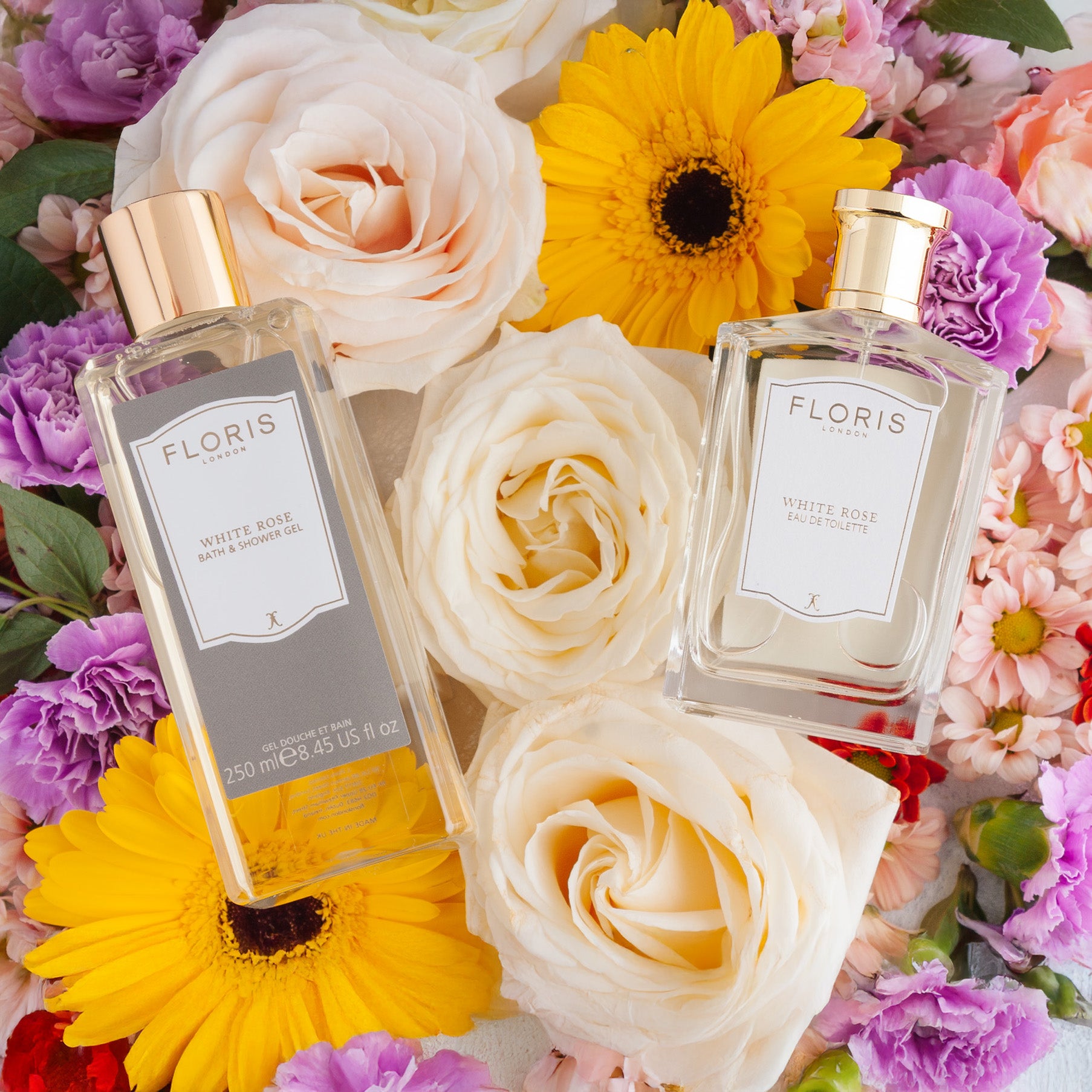Two Floris London White Rose Mother's Day Pairing bottles on white, yellow, and purple flowers, including roses and gerbera daisies.