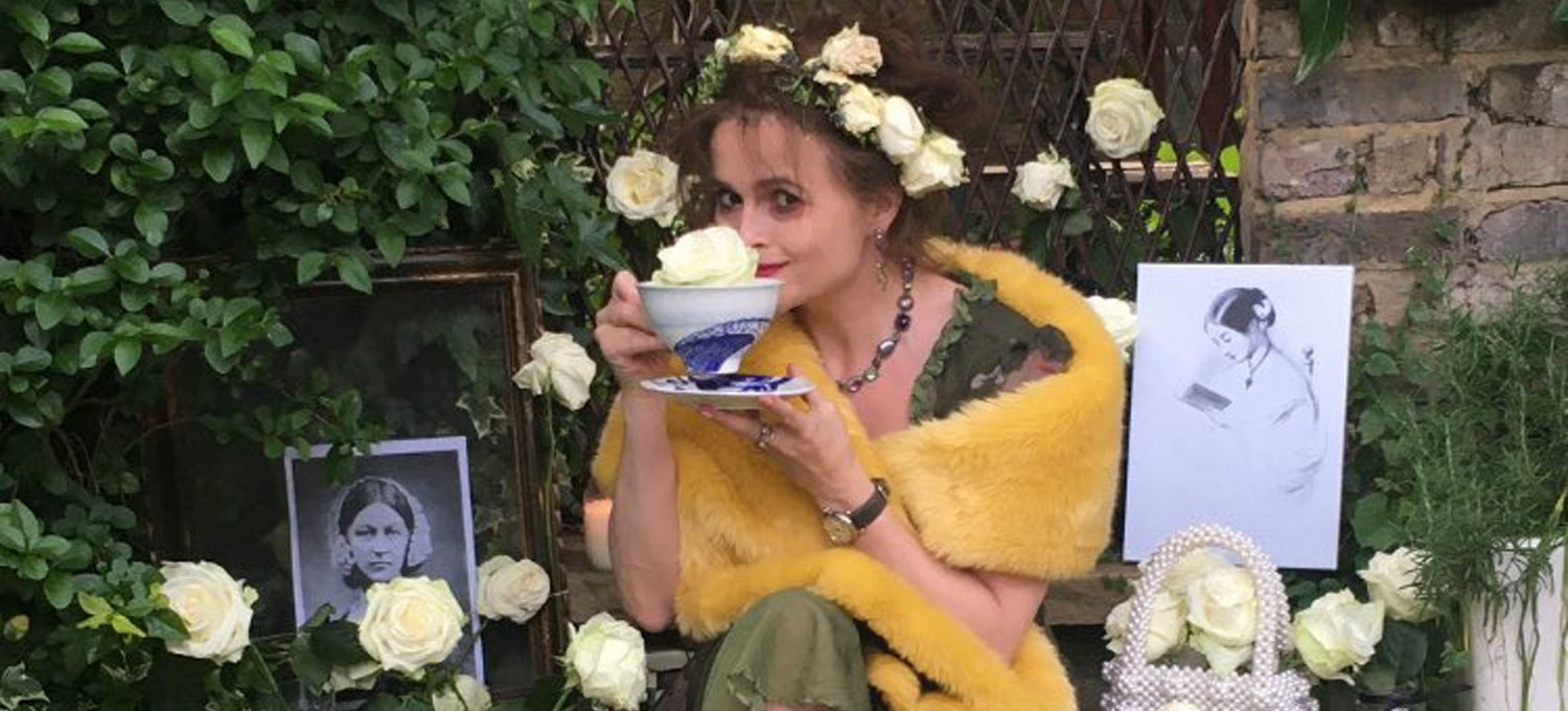 Person with a flower crown holding a teacup, surrounded by white roses and black-and-white photos near a brick wall.