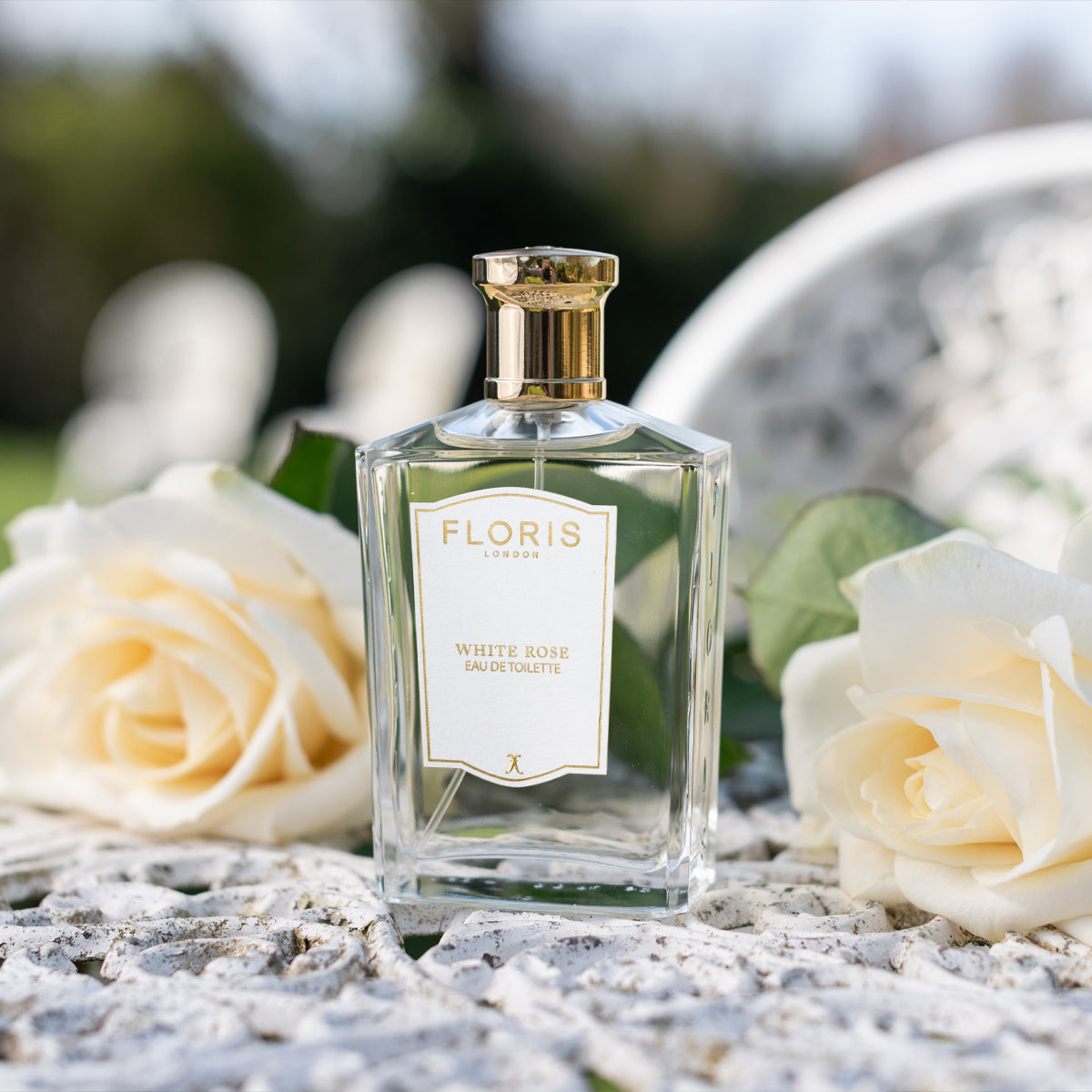 Floris London White Rose Eau de Toilette bottle beside velvet roses on a patterned surface, exuding hints of warm amber.