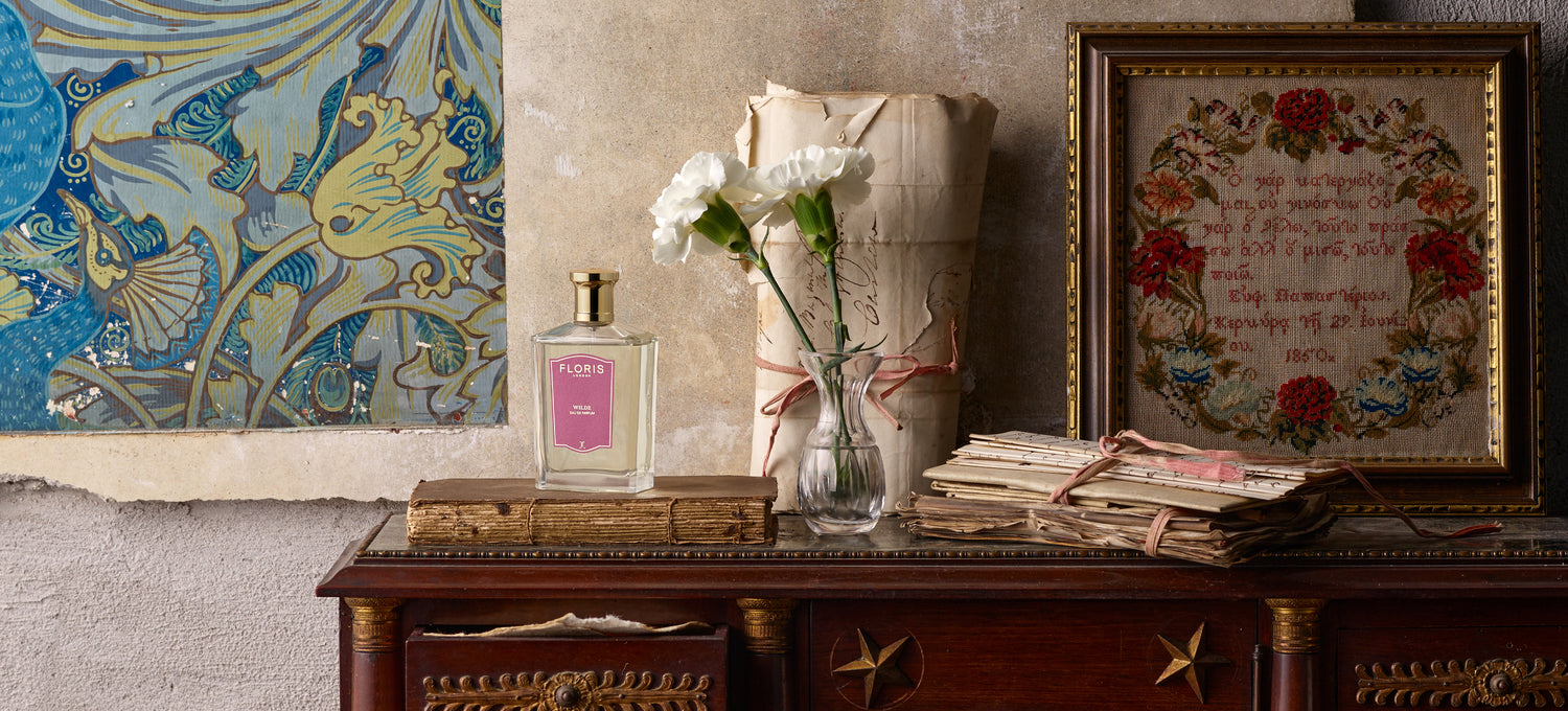 Vintage table with flowers in a vase, perfume bottle, old books, letters, and framed art against a worn wall.