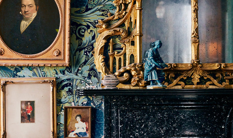 A room with ornate wallpaper, framed portraits, a gilded mirror, and a small statue on a black marble fireplace mantle.