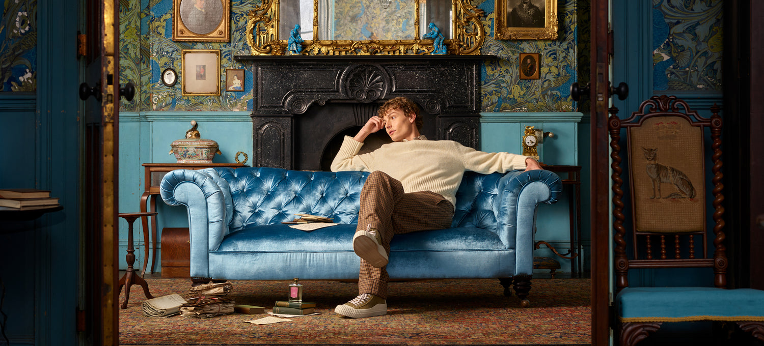 A person lounging on a blue velvet sofa in a vintage-style room with ornate wallpaper and framed pictures.