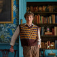 A person in a patterned sweater vest stands by a bookshelf, embodying the individuality of Floris London's Wilde amidst patterned wallpaper and an Oscar Wilde-like portrait.