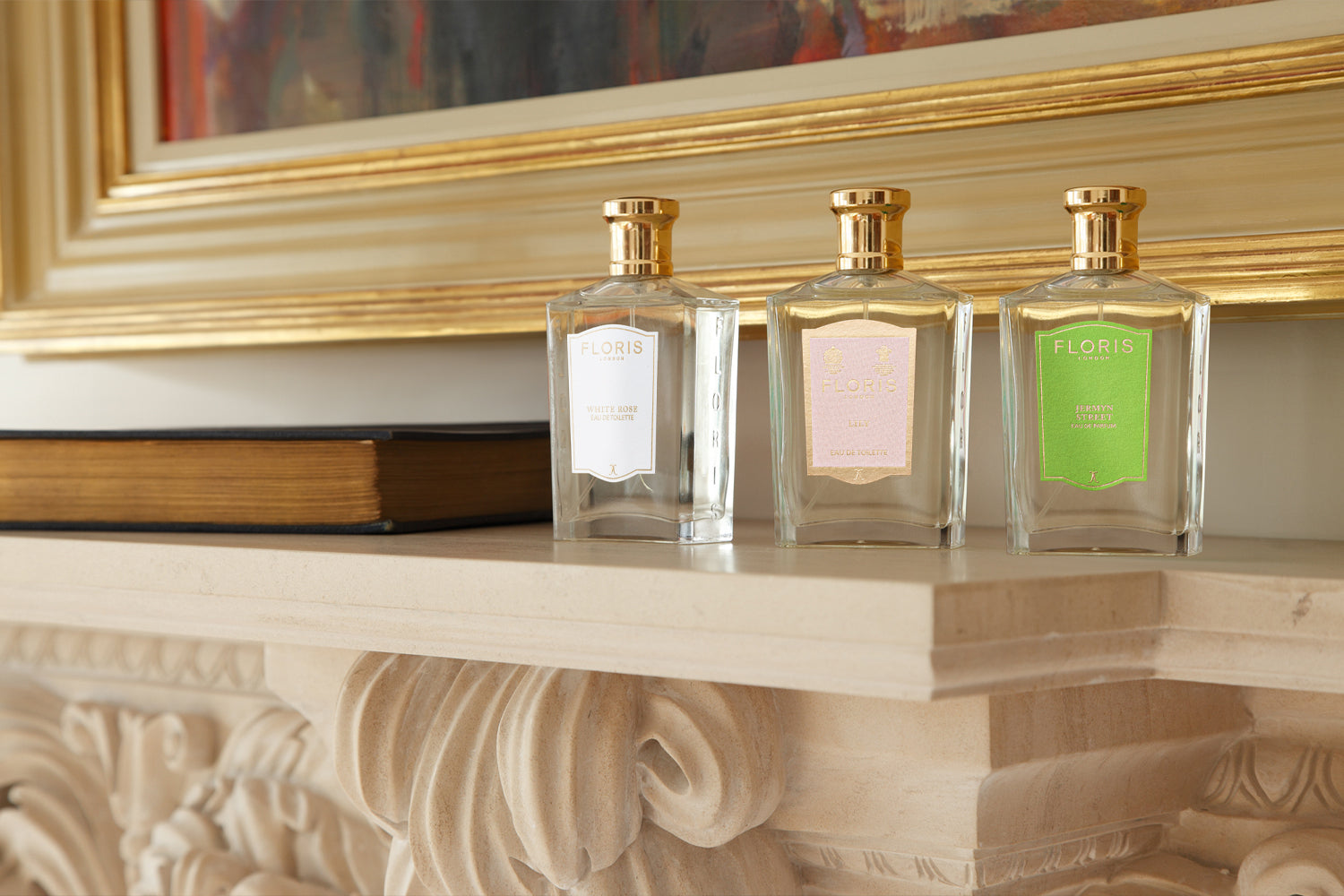 Three Floris perfume bottles in different scents on a decorative mantelpiece with a book and framed artwork above.