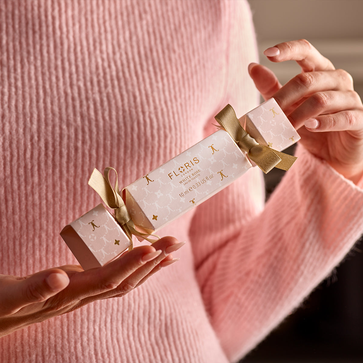 A White Rose Christmas Cracker from Floris London, being held in front of a person with a wool, pink jumper. This luxurious gift is perfect for the holiday season.